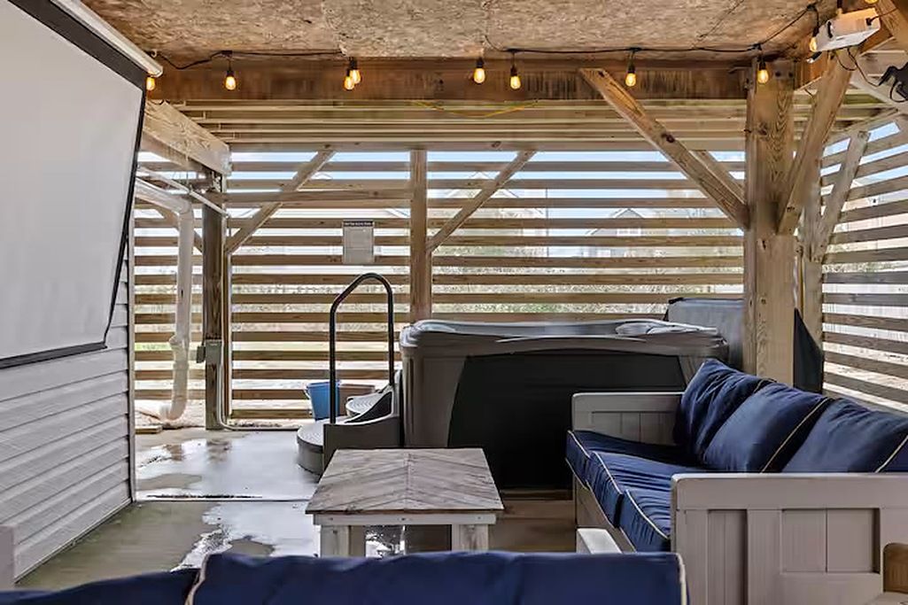 A covered outdoor seating area with a hot tub, wooden beams, and string lights.