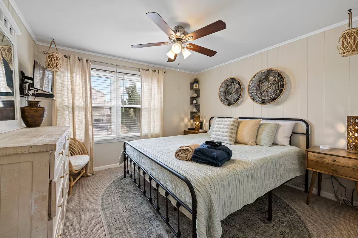 A bedroom with a bed, a ceiling fan, a window with curtains, a bedside table, and wall decor.