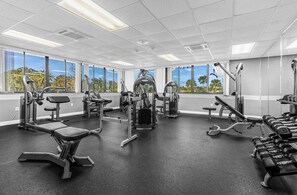 Un gimnasio con diversos equipos de ejercicio, amplias ventanas y vistas a palmeras.