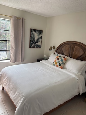 Una cama bien hecha con ropa blanca, un cabecero de madera, una lámpara de pared y un cuadro con una palmera.