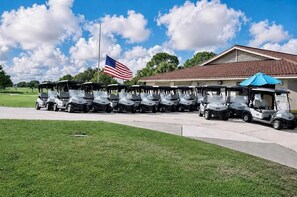 Une rangée de voiturettes de golf est stationnée devant un bâtiment orné d’un mât et d’un drapeau américain.