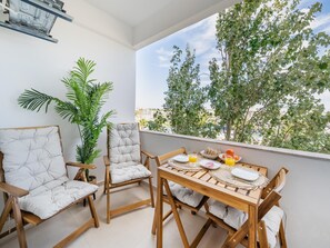 A balcony with a wooden table set for breakfast, two upholstered chairs, a potted plant, and a view of trees and buildings. - Stylish Central Studio with Balcony, Lagos (Lagos)