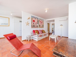 Ein geräumiger Wohnbereich mit einem roten Lounge-Sessel, einem weißen Sofa mit bunten Kissen und einem großen Gemälde an der Wand.