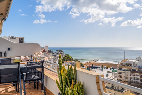 Uma varanda com vista para o mar, uma mesa com cadeiras e um vaso de planta.