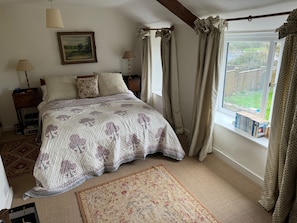 A bedroom with a bed, a nightstand, a lamp, a window with curtains, and a view of the outdoors. - Cottage Sleeps 7 with Garden & Free Parking (Dorchester)