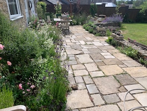 A stone pathway leading to a patio area with a table and chairs, surrounded by lush greenery and flowering plants. - Cottage Sleeps 7 with Garden & Free Parking (Dorchester)