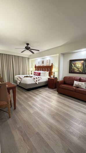 A hotel room with a bed, a wooden floor, a brown leather sofa, a wooden side table, and a ceiling fan. - Cozy 2-bedroom Apartment With Shared Pool in Orlando (Orlando)
