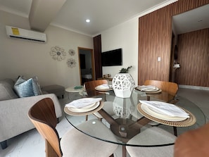 A modern living room with a glass-top dining table, wooden chairs, a sofa, and a flat-screen TV mounted on the wall. - Vrbo Property (Santo Domingo)