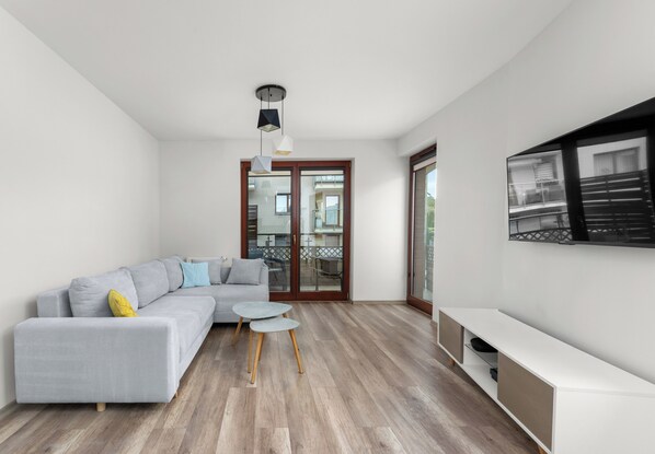 Een moderne woonkamer met een grijze hoekbank, een klein rond tafeltje en een flatscreen tv aan de muur.
