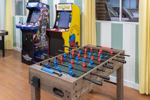 Ein Holztischkicker mit roten und blauen Spielfiguren vor einer gestreiften Wand, im Hintergrund sind Spielautomaten zu sehen.