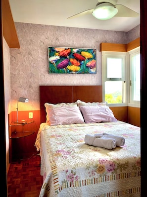 A hotel room with a bed, a painting above it, a nightstand, a lamp, and a window with curtains. - Vrbo Property (Gramado)