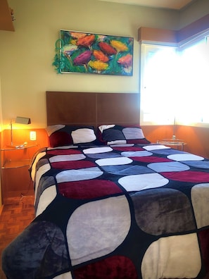 A bedroom with a large bed, a nightstand, a lamp, a painting, and a window. - Vrbo Property (Gramado)