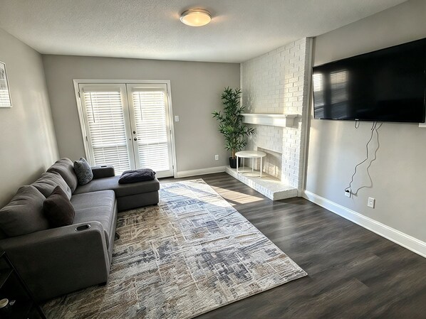 A living room with a sectional sofa, a patterned rug, a flat-screen TV mounted on the wall, and a potted plant. - The Modern Stay - Near Stadiums & DFW Airport (Addison)