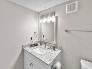 A bathroom with a granite countertop sink, a mirror with lights, a towel rack, and a toilet paper holder. - The Modern Stay - Near Stadiums & DFW Airport (Addison)