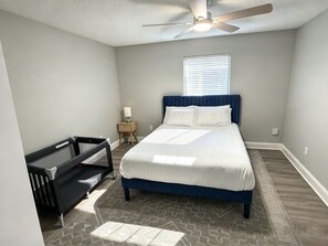 A bedroom with a bed, a ceiling fan, a window with blinds, a bedside table with a lamp, and a crib. - The Modern Stay - Near Stadiums & DFW Airport (Addison)