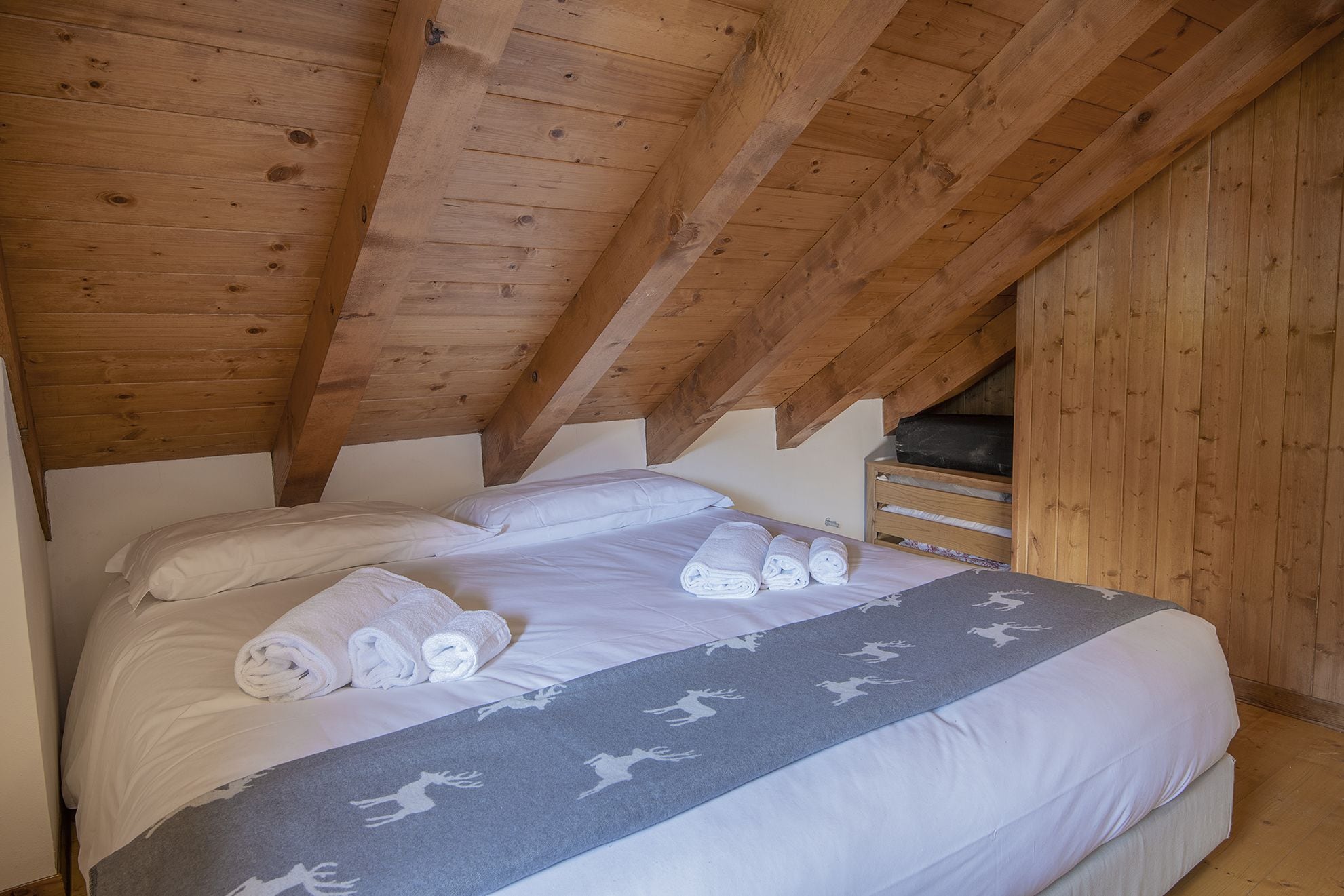 Ein Schlafzimmer mit Holzbalkendecke, einem Bett mit weißen Leinen und einer grauen Decke mit Rentier-Motiven.