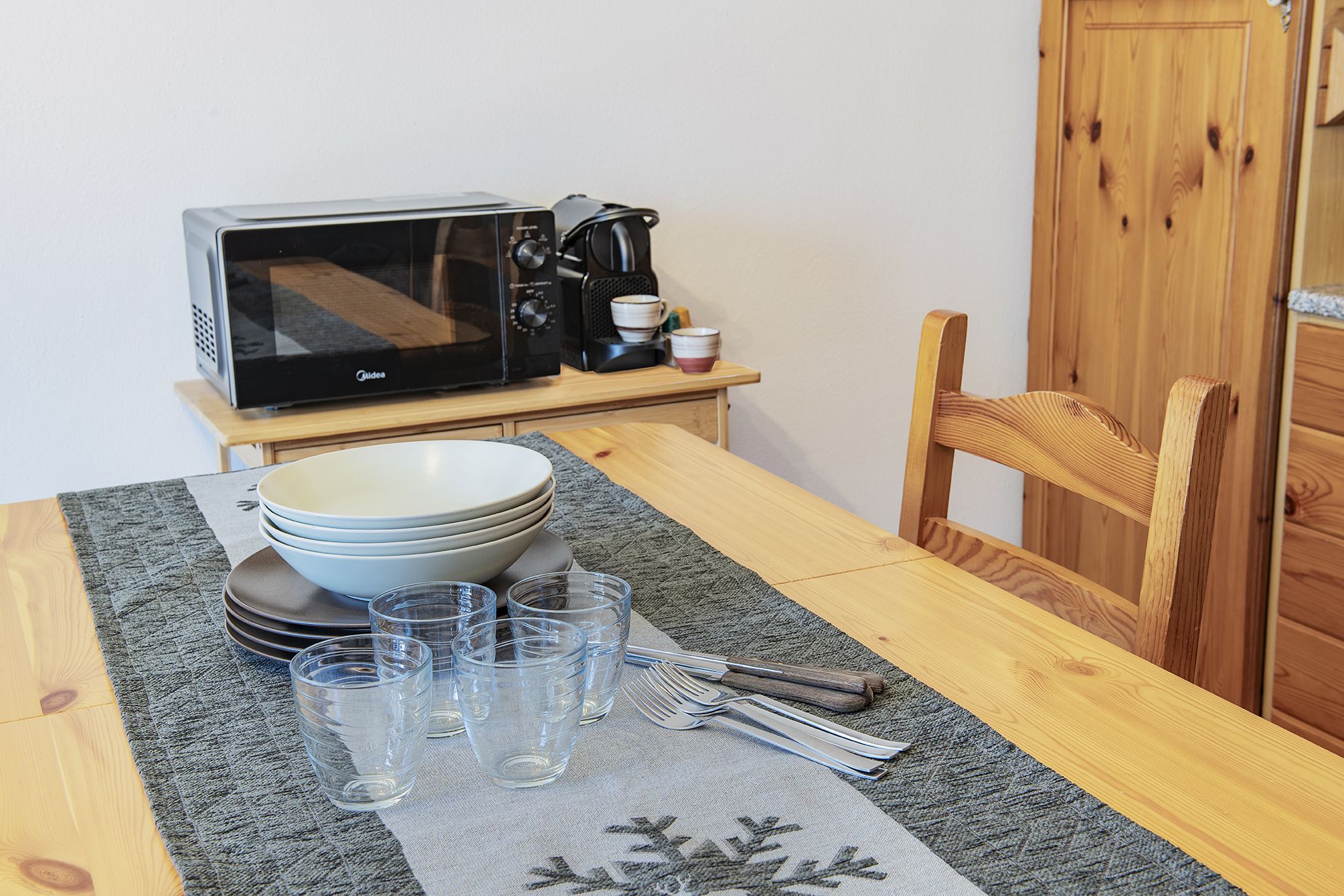 A dining table with a microwave, a coffee maker, bowls, glasses, and cutlery.