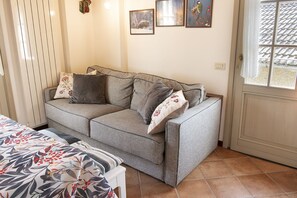 Habitació amb un sofà gris, un llençol estampat i una finestra amb vista a un teulat llostionat.