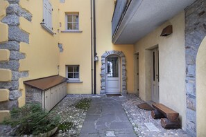 A narrow alleyway with a stone wall on one side and a yellow building on the other. There is a wooden bench and a door with a stone archway. - AffittiPontediLegno - Sguardo sulle cime (Ponte di legno)