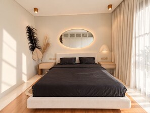 Een moderne slaapkamer met een groot bed, nachtkastjes, een ronde spiegel en uitzicht op het groen.