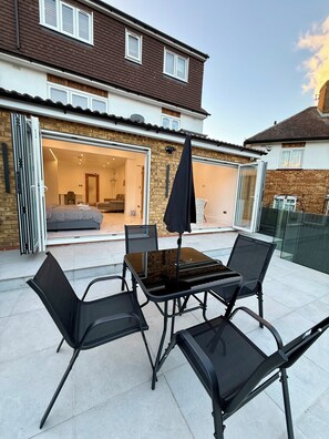 A patio with a glass table and chairs, a black umbrella, and a view of a house with a brick facade and a garage door. - Modern, Luxury Stay with Free Parking for 3 car's easy access to central London (Potters Bar)