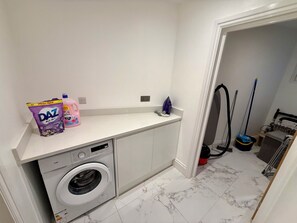 A laundry area with a washing machine, a shelf with cleaning products, and a mop. - Modern, Luxury Stay with Free Parking for 3 car's easy access to central London (Potters Bar)