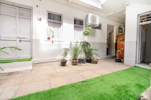Un pati amb sòl de pedra, plantes en testos, una zona verda i un edifici amb portes i finestres blanques.