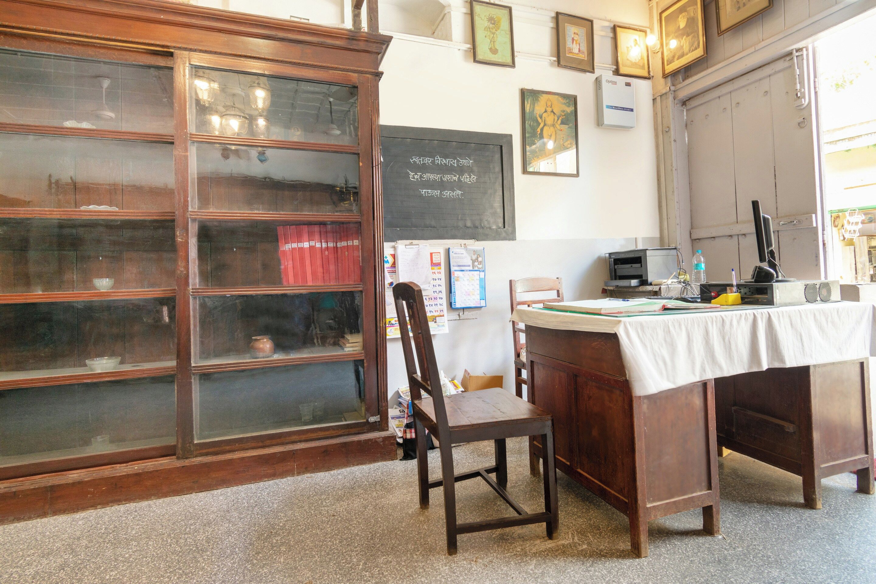 A room with a wooden cabinet with glass doors, a desk with a computer, and a chair. There are framed pictures on the wall and a blackboard with text.