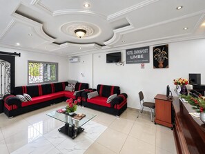 Un hall d’hôtel comprenant un comptoir d’accueil, deux canapés noirs avec des coussins rouges, une table basse en verre et un plafond doté d’un éclairage encastré.