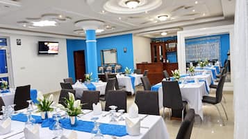 Un comedor de hotel con mesas dispuestas para el servicio, una zona de bar y una pantalla de televisión.