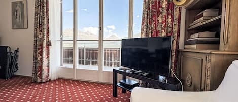 A hotel room with a large window, a flat-screen TV, a bookshelf, and a red patterned carpet.