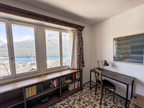 A room with a large window, a bookshelf, a desk with a lamp, and a patterned carpet. - Fullun 14 by Interhome (St. Moritz)