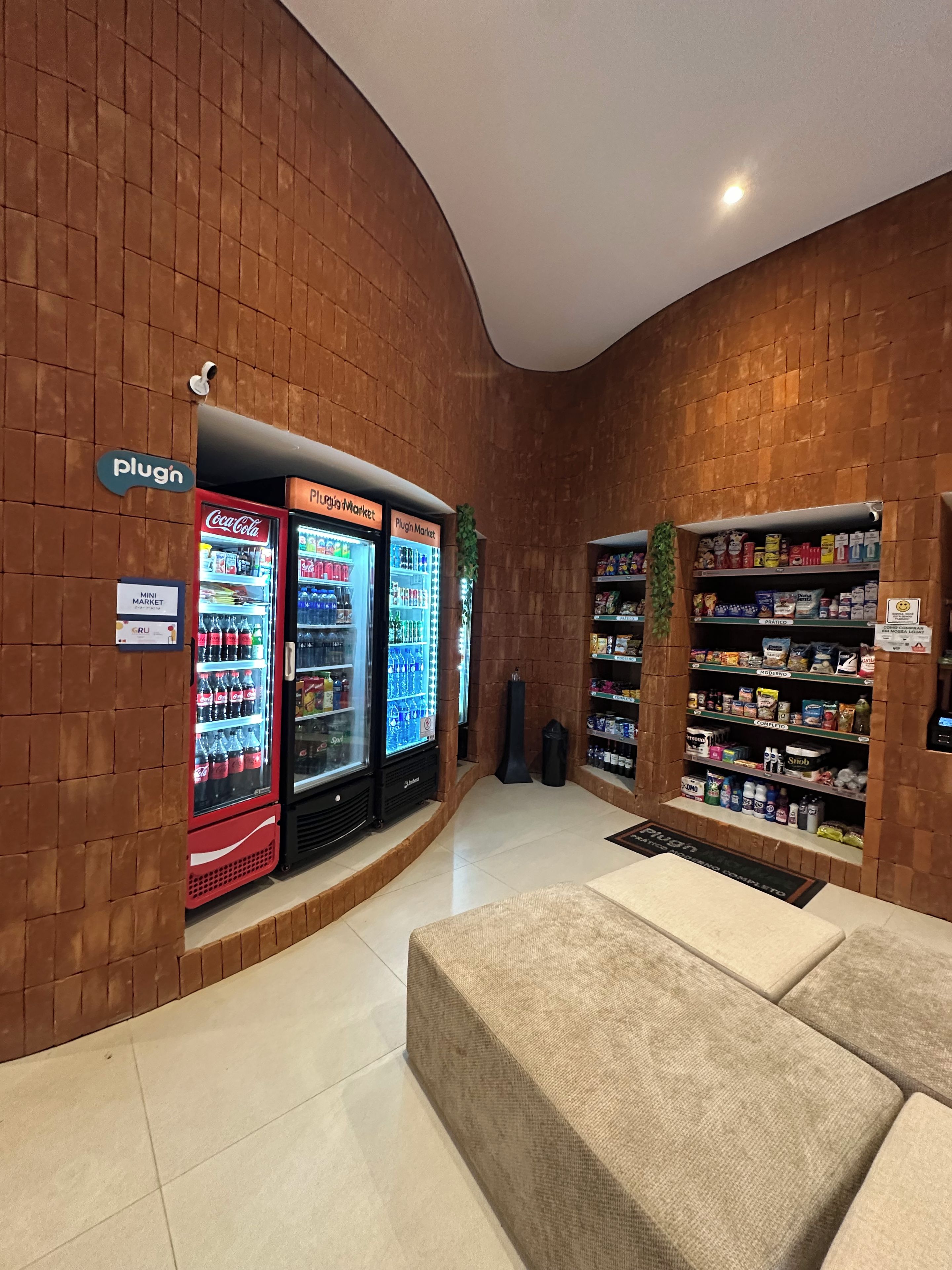 A lobby with a curved brick wall, a Coca-Cola refrigerator, a Plugio Market refrigerator, and a snack shelf.