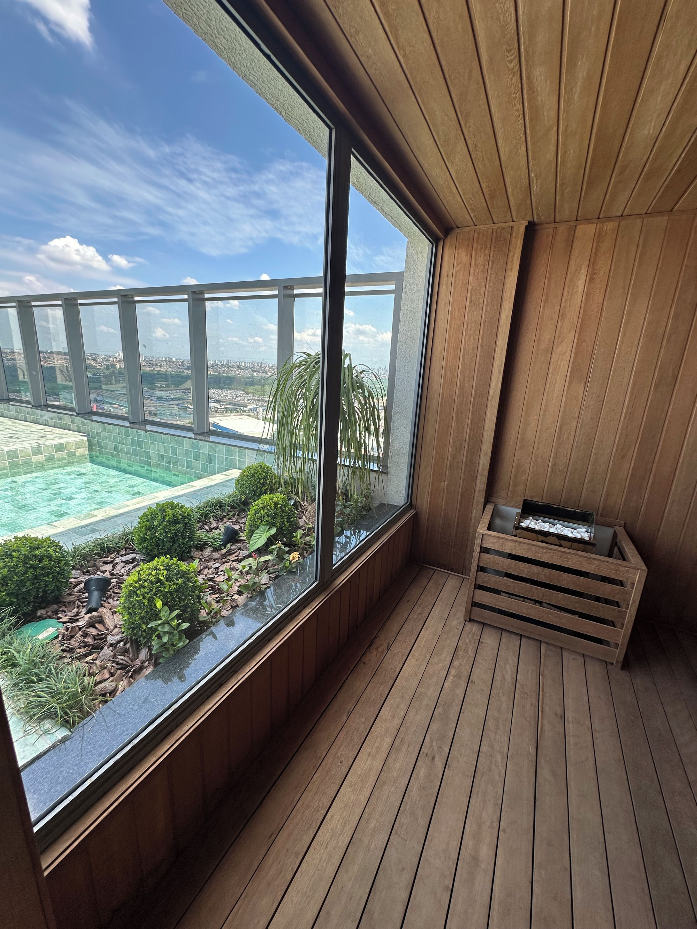 A balcony with a view of a cityscape, wooden flooring, and a small seating area with cushions.