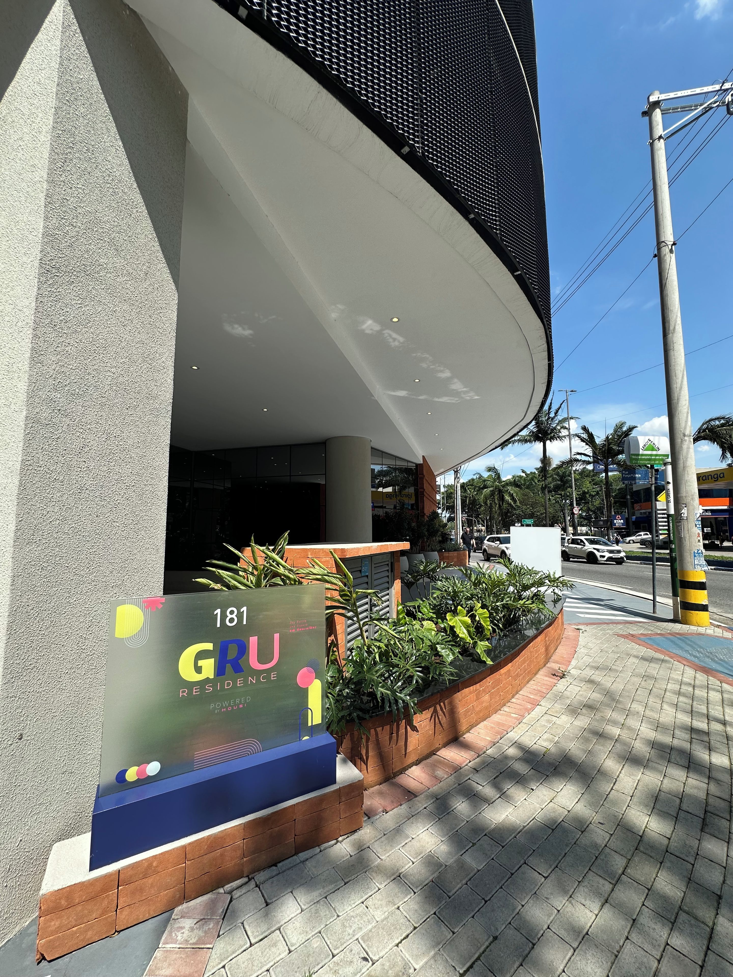 A modern building entrance with a sign for '181 GRU RESIDENCE' and a landscaped area with plants.