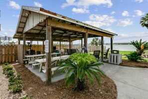 A covered outdoor seating area with picnic tables and a grill, overlooking a body of water. - Always 5 O'clock - Lagoon Front, Private Beach Access, Pool & Kayaks (Gulf Shores)