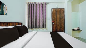 Habitación de hotel con cama, puerta de madera, ventana con cortinas estampadas, equipo de aire acondicionado montado en la pared y una pequeña estantería con tetera y tazas.