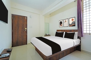 A hotel room with a bed, a bedside table with a remote control, a wall-mounted TV, and a window with a curtain. - Collection O Virajpete Bus Terminal (Coorg)