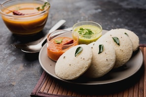 A plate with idlis and three different chutneys, a bowl of soup, and a spoon on a wooden surface. - Collection O Virajpete Bus Terminal (Virajpet)