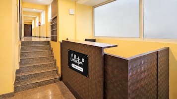 Zona de recepción de un hotel, con mostrador, escalera y un pasillo que lleva a una puerta.