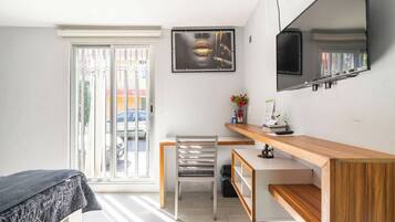 Un dormitorio con cama, escritorio, silla, ventana con vista a automóviles y televisor montado en la pared.