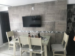 A modern dining area with a flat-screen TV mounted on the wall, a small table with a laptop, and two chairs.