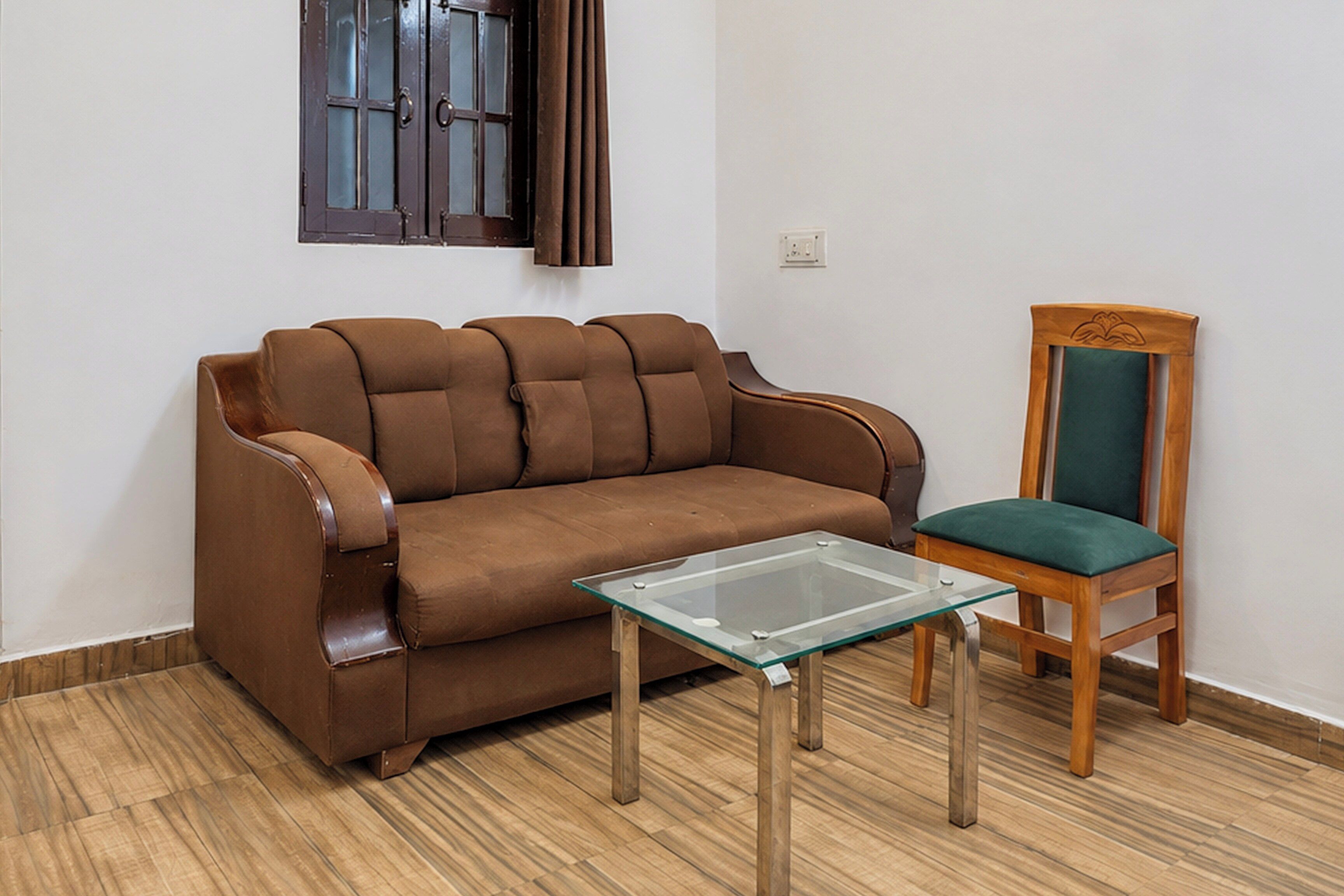 A brown sofa, a glass-top coffee table, and a wooden chair with a green cushion in a room with wooden flooring and a white wall.