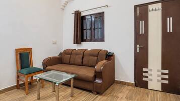 Una habitación con un sofá marrón, una mesa de vidrio, una silla de madera con cojín verde, una ventana con cortinas y una puerta con picaporte.