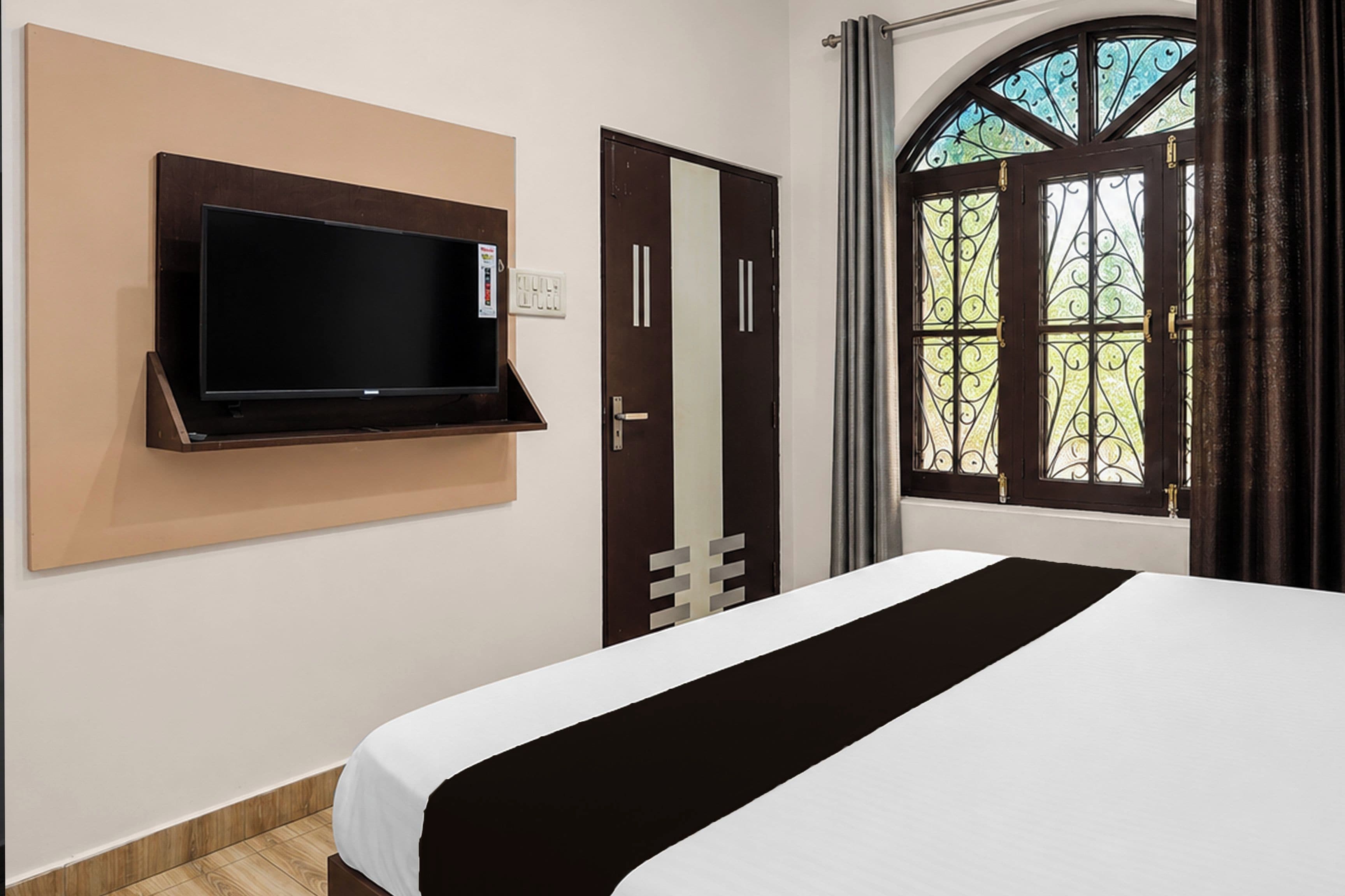 A hotel room with a bed, a mounted TV, a door with a mirror, and a window with curtains.