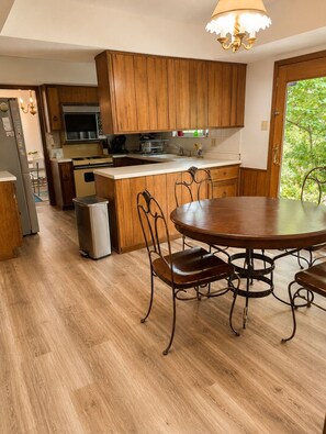 Una cocina con armarios de madera, un refrigerador de acero inoxidable y una zona de comedor con una mesa redonda y sillas metálicas.