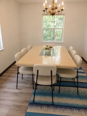 Una sala menjador amb una taula rectangular, sis cadires, un penjador de llum, una finestra amb vista a arbres i una catifes amb dibuixos.