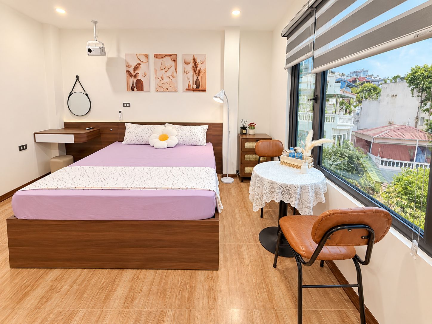 A modern bedroom with a large bed, a bedside table, a chair, a small table with a lace tablecloth, and a view of the city through a large window.