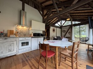 A rustic kitchen with wooden beams, a white dining table with chairs, and a view through large windows. - Les Pins Vacation Rental - CAME - Vacation Rental (CAME)