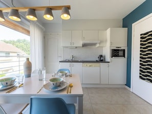 A modern kitchen with white cabinets, a dining table set with plates, glasses, and cutlery, and a view of the outdoors through a window. - Lovely apartment for 3 guests with pool, WIFI, TV and balcony (De Haan)